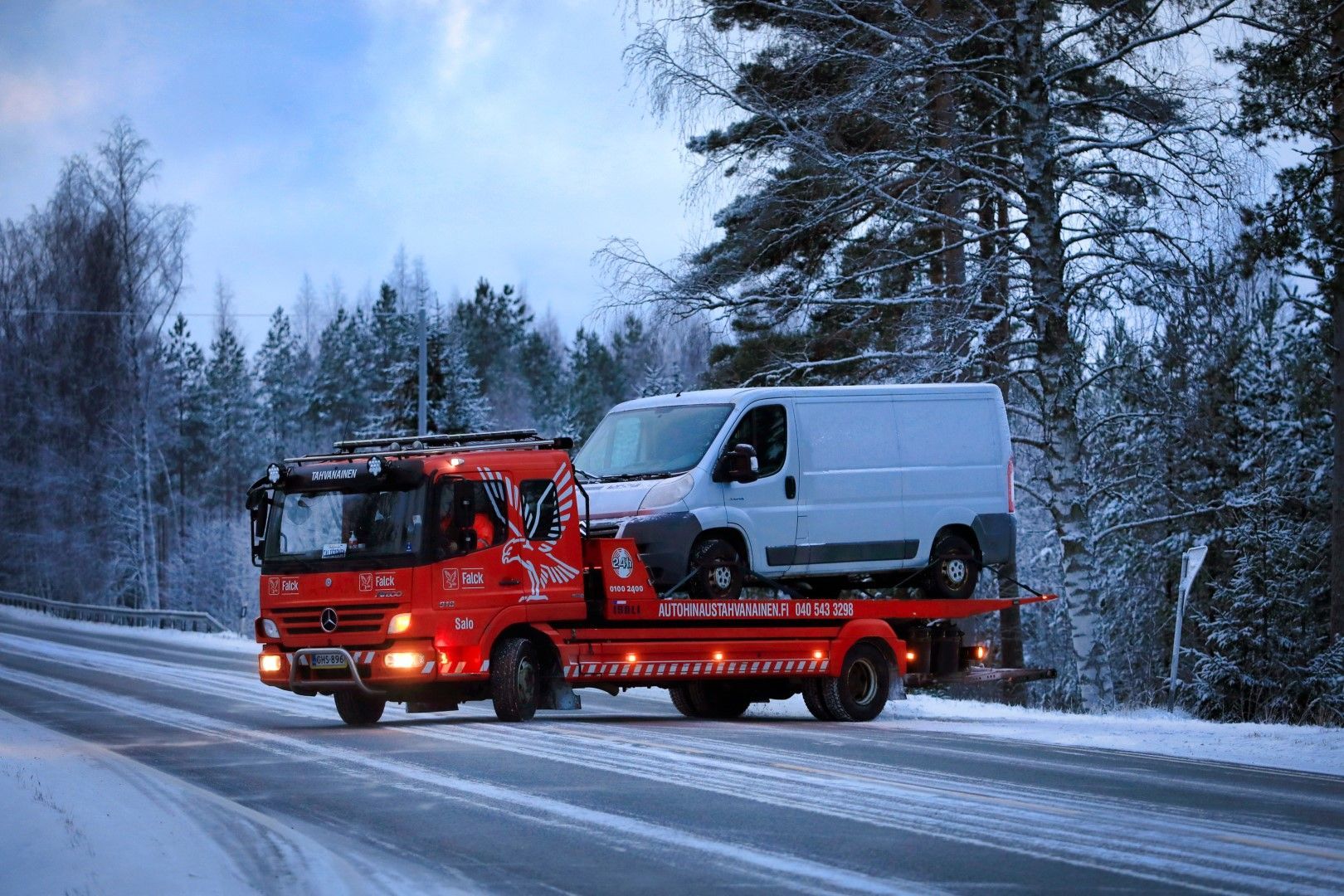 A tow truck is carrying two vans down a snowy road.